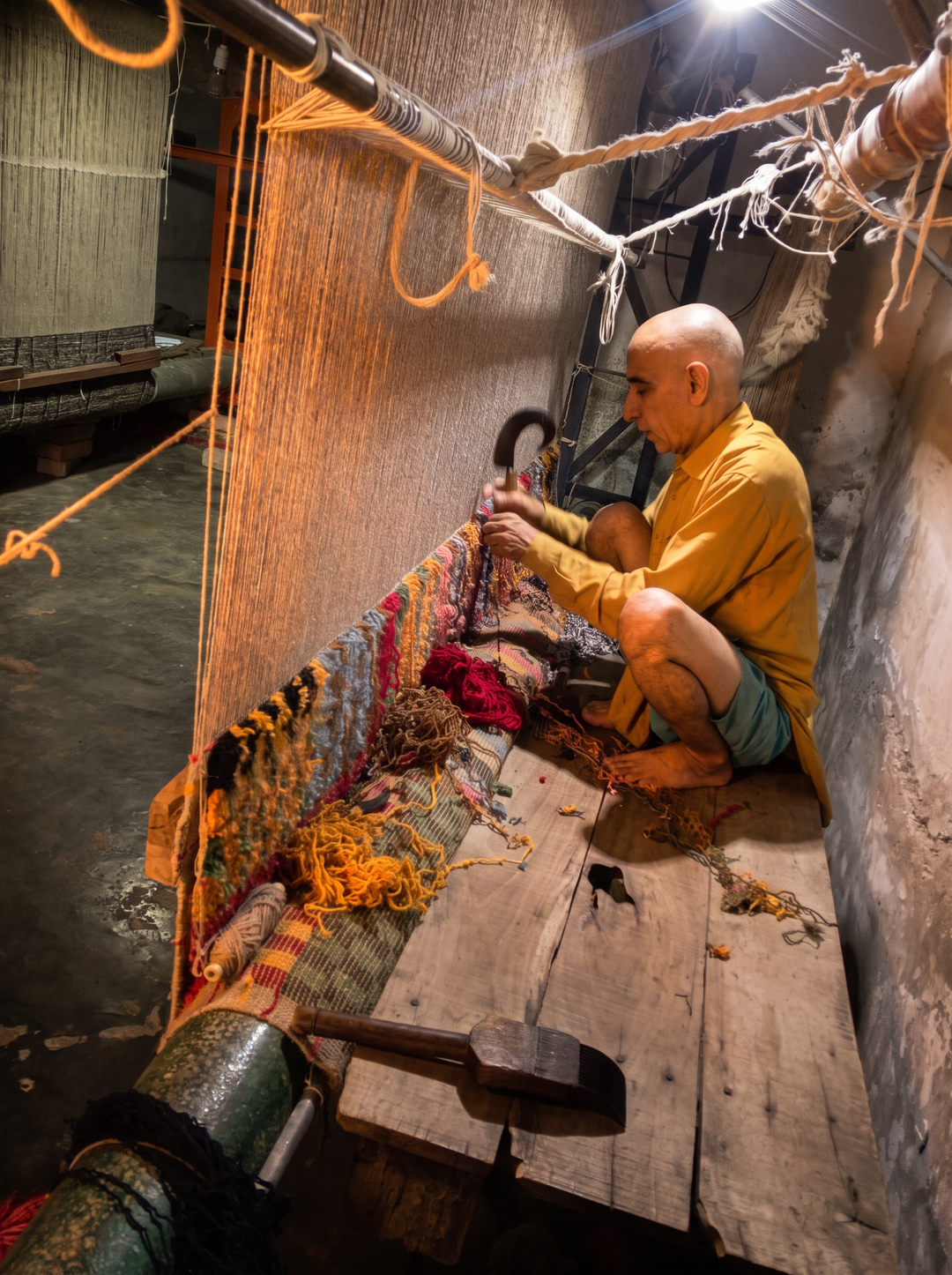 A weaver hand-knotting colored wool onto a loom, using a hooked tool to tie each knot at an Imam Carpets workshop